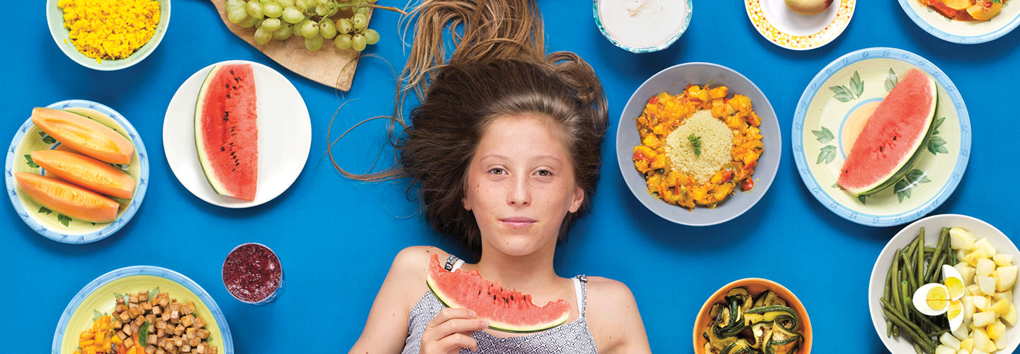 A young girl eats a slice of watermelon with dishes of food around her head.