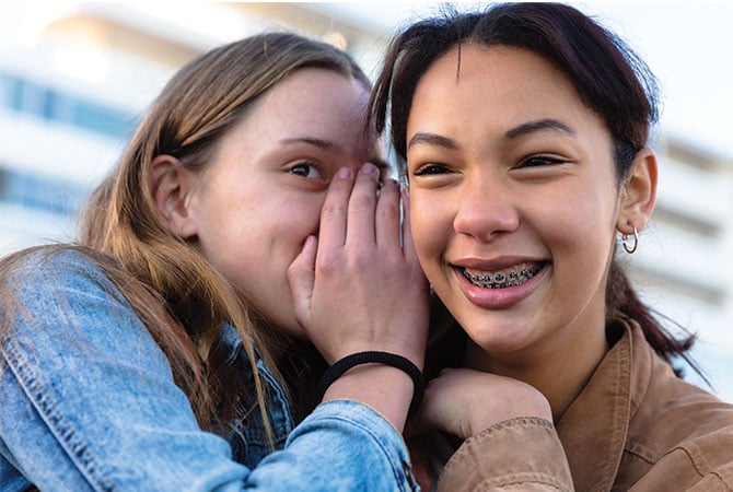 A teen whispering into the ear of another teen