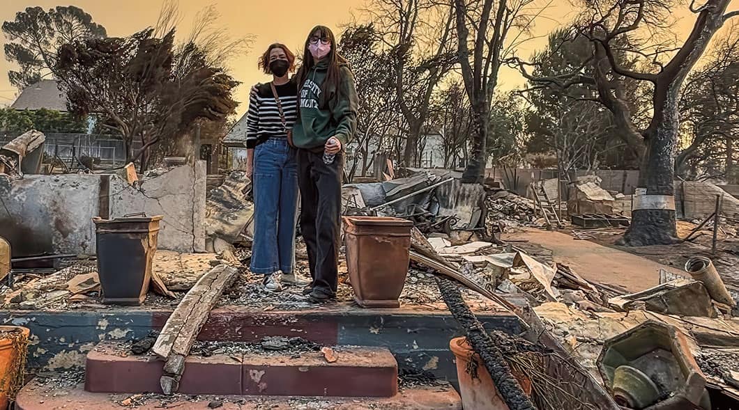 Two people stand amid fire-damaged home ruins under an orange sky