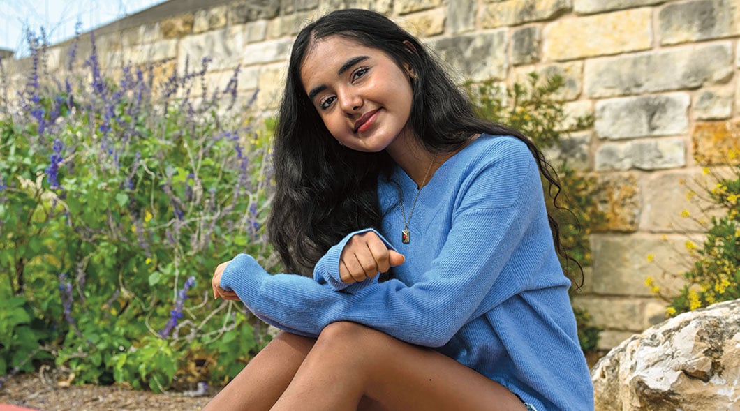 A girl in a blue sweater sits outdoors near a stone wall with purple flowers
