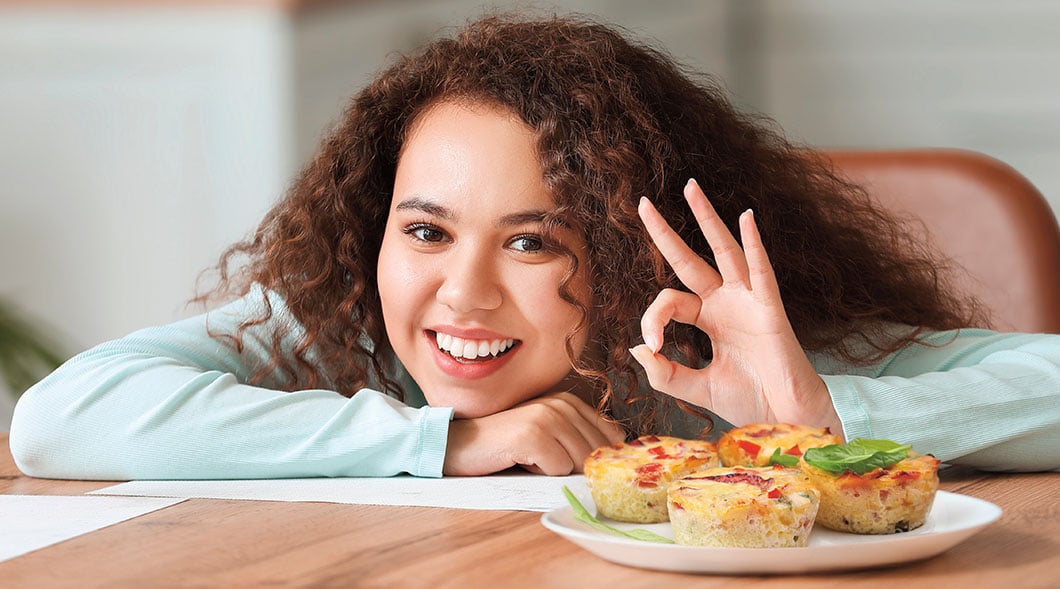 A girl leans on a table and makes an okay sign next to a plate of mini egg muffins