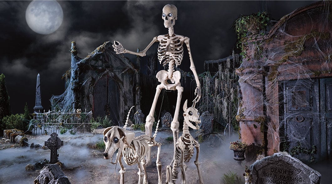 A large skeleton figure walks two skeleton dogs through a foggy graveyard at night