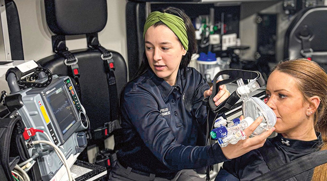 A paramedic holds a breathing mask toward a woman inside an ambulance with a cardiac monitor nearby