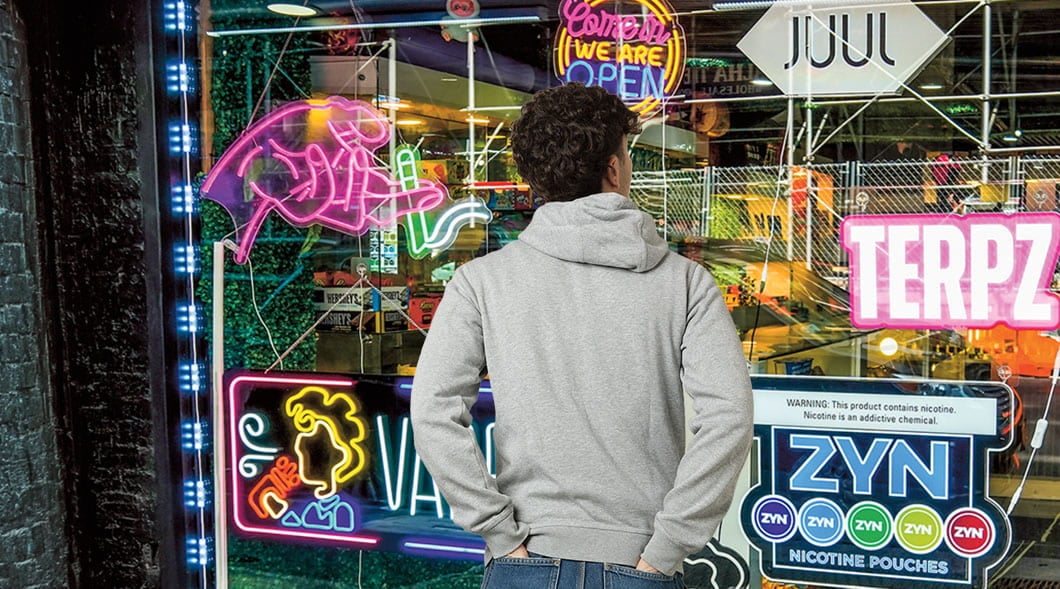 A person stands outside a vape shop window with neon signs and Juul and Zyn nicotine product ads