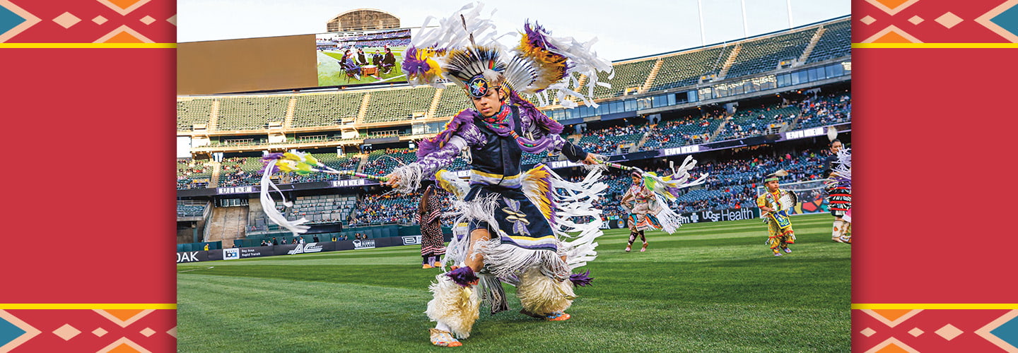A dancer in traditional regalia performs on a stadium field before a crowd of spectators
