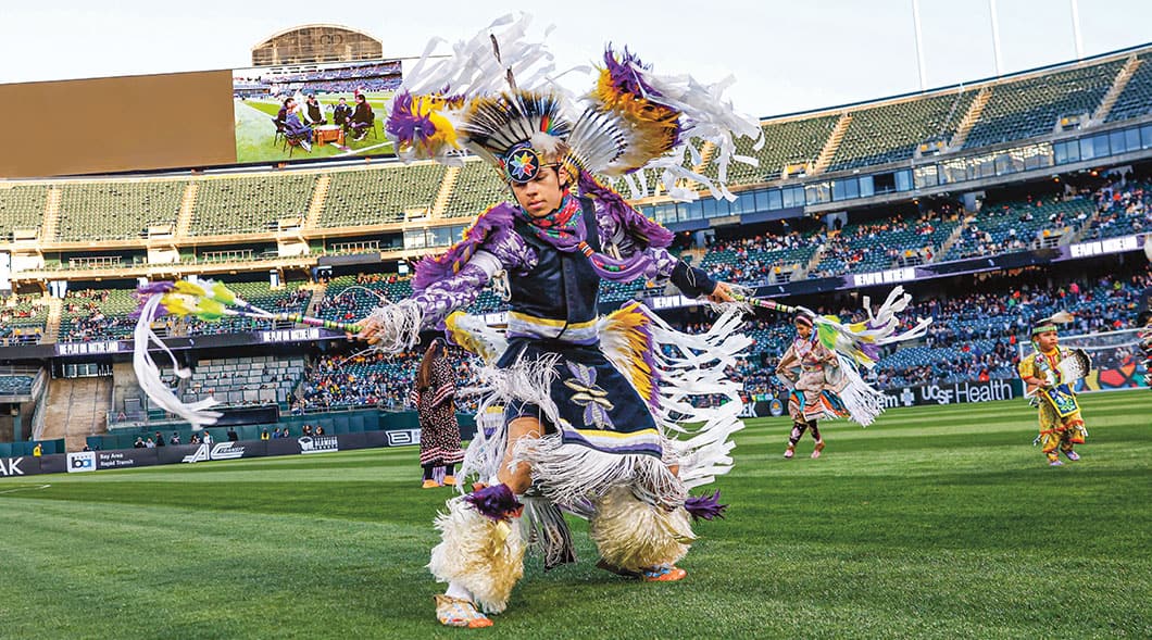 A dancer in traditional regalia performs on a stadium field alongside other dancers