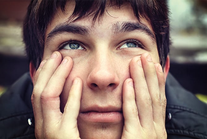 A teenage boy looking up with both hands on his cheeks.